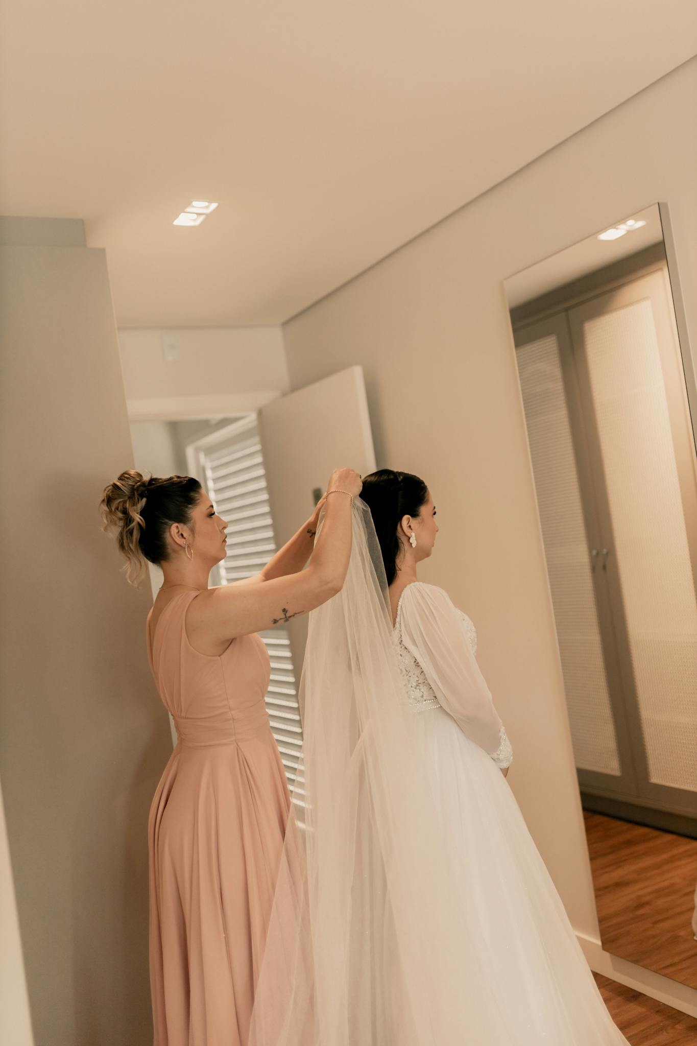 Bridesmaid in peach dress helps bride with veil indoors.